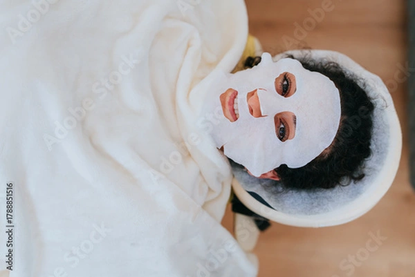 Fototapeta A serene facial treatment scene where a person rests under a soft blanket, wearing a revitalizing sheet mask in a tranquil spa.