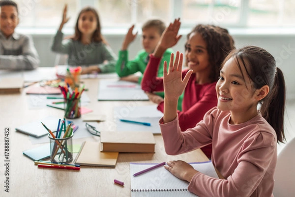 Fototapeta Multiethnic school kids sit at their desks in a bright classroom, eagerly raising their hands to answer questions. They smile, showing excitement and enthusiasm for learning.