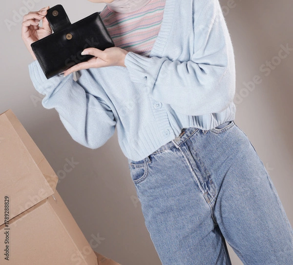 Fototapeta Young woman's body in a knitted sweater and jeans and her hand showing an elegant shiny black leather purse