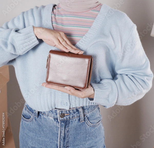 Fototapeta Young woman's body in a knitted sweater and jeans and her hand showing an elegant shiny brown leather purse
