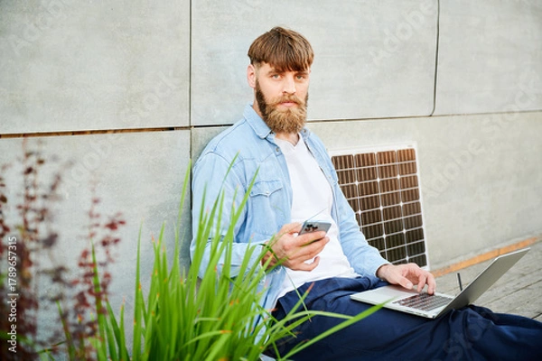 Fototapeta Bearded man works on laptop on lap and smartphone. Solar panel nearby, emphasizing sustainable, modern eco-friendly workspace that combines technology and sustainability.