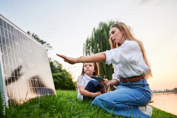 Fototapeta Mother connecting smartphone to photovoltaic solar panel for charging, explains its workings to daughter, sit on grass by lake. Integration of sustainable renewable energy into everyday life.