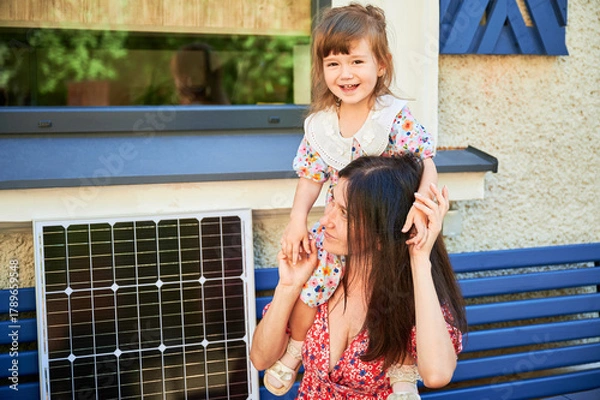 Fototapeta Mother sits on bench with her young daughter, both smiling joyfully. Solar panel placed beside them, symbolizing embrace of renewable energy and bright, sustainable future for next generation.