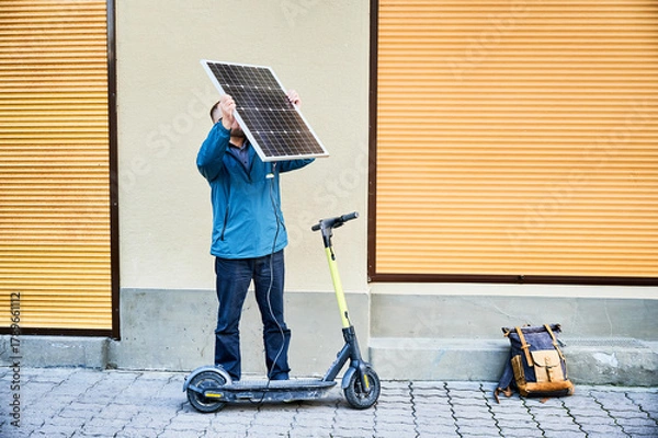 Fototapeta Male person holds photovoltaic solar panel, for charging electric scooter in urban settings. Integration of solar power as sustainable energy source, promoting eco-friendly urban transportation.