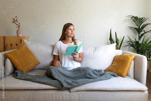 Obraz Woman reading at home drinking cup of coffee looking away. Female relaxing at home reading book enjoying herbal tea.