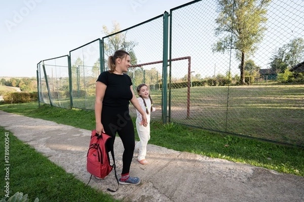 Fototapeta Mother and daughter stroll along a path beside a fenced field, sharing a moment of connection on a sunny day.