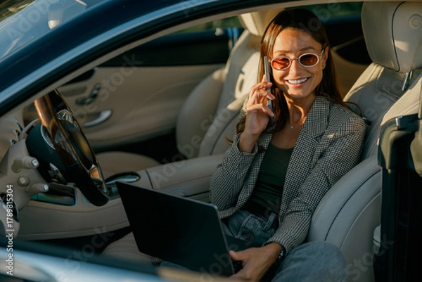 Fototapeta Woman working on laptop inside car, making playful peace sign during video call while managing schedule, blazer-clad professional