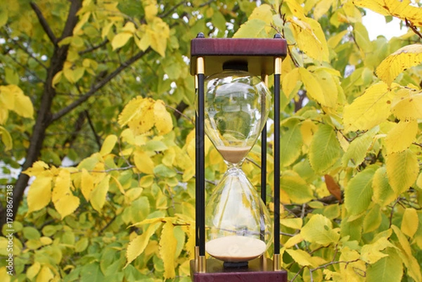 Obraz An hourglass against the backdrop of a tree with yellow leaves. The hourglass symbolizes time, balance, cyclicality, and transience.