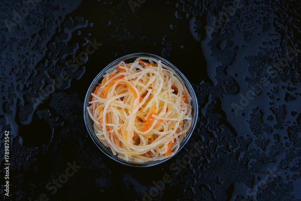 Fototapeta Glass noodle salad with carrots in a glass bowl on a black background with water drops. View from above.