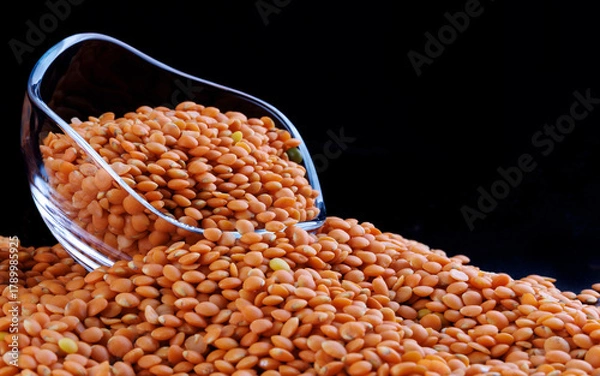 Fototapeta Red lentils in a transparent glass cup on a black background. Side view. Lots of lentils around the cup.