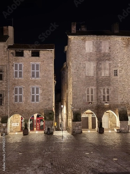 Fototapeta Illuminated medieval square with stone buildings and arched entrances at night