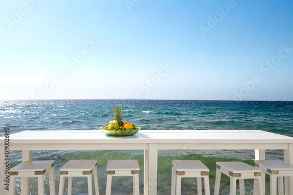 Fototapeta white wooden table with empty chairs on seascape backdrop under clear blue sky on sunny summer day. plate of fruit