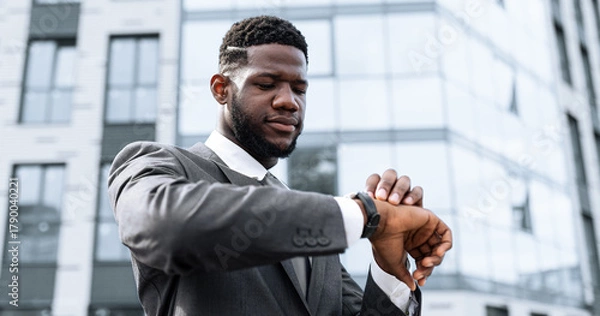 Fototapeta A man dressed in a formal suit is checking his watch while standing in front of a contemporary office building. The scene captures a moment of waiting or planning in an urban environment.
