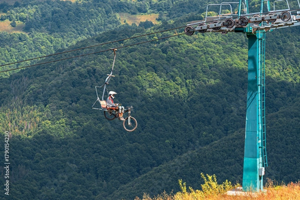 Fototapeta A mountain biker and their bicycle ascend a heavily forested mountain range via a chairlift on a bright, sunny day.