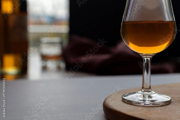 Fototapeta Elegant glass of amber brandy or cognac on a wooden stand in a dim, atmospheric bar or tasting room. A bottle and blurred background create a sophisticated mood