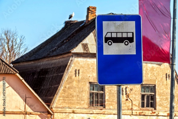 Fototapeta Blank bus stop sign near an old building with a stork resting on the roof in daylight