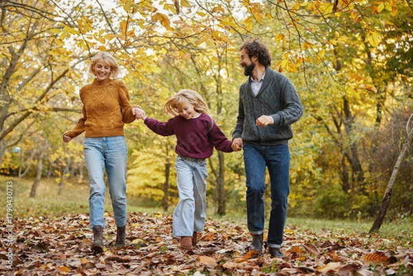 Obraz A family of three walks hand in hand through a vibrant park filled with colorful autumn leaves. They smile and share joyful moments on a sunny weekend.