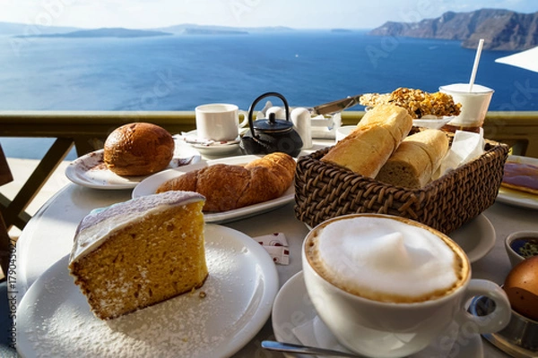 Fototapeta Beautiful island breakfast with Aegean sea view including cappuccino cup, cake, baguette, croissant, boiled egg, hot tea and chocolate brioche bun on white marble table