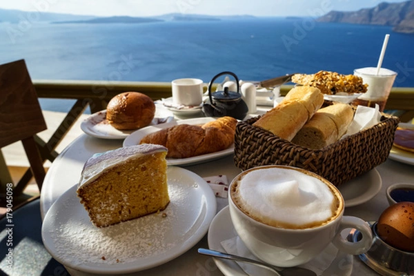 Fototapeta Enjoy island breakfast with beautiful Aegean sea view and morning sunlight including cappuccino cup, cake, baguette, croissant, boiled egg, hot tea and chocolate brioche bun on white marble table