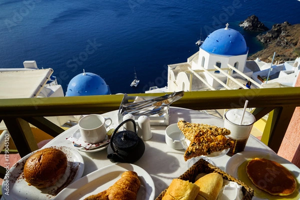 Fototapeta Breakfast with beautiful Aegean sea view, boat and Oia church including pancake, baguette, croissant, hot tea, chocolate brioche bun and fruity cereal bar on white marble table