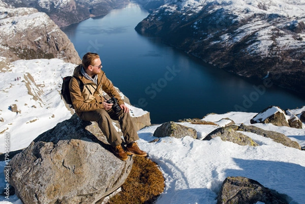 Fototapeta Man with Camera on Snowy Cliff Overlooking Fjord