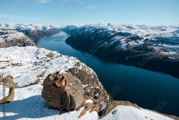 Fototapeta Man on Snowy Cliff Overlooking Fjord and Mountains