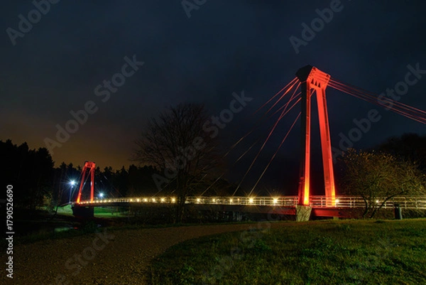 Obraz Night view of an illuminated pedestrian suspension bridge with red lights, glowing against a dark cloudy sky and forest background.
