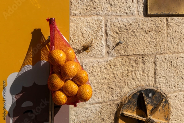 Fototapeta A mesh bag of bright oranges hangs against a vivid yellow wall with a graphic of an old person's face, beside a textured stone wall.