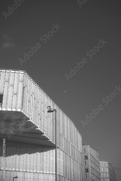 Fototapeta Oslo, Norway, translucent, ribbed modern blocks rise above a lone street lamp, light and shadow grid the facade, with a faint daytime moon in a clear minimalist sky overhead calm