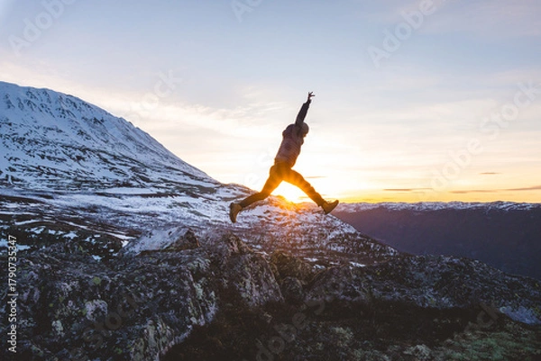 Obraz Traveler jumps from rocks at sunset near Gaustatoppen Mountain in Norway. Golden light, snow peaks, and open sky capture pure joy and freedom of adventure