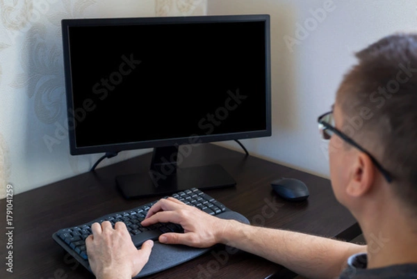Fototapeta Remote work from home. Young man wearing glasses is focused on computer with black screen in stylish home office, featuring wooden desk and minimalistic decor, creating productive work environment