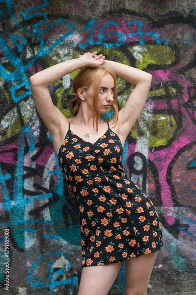 Obraz Blonde sensual woman with long legs posing confidently in front of a colorful graffiti wall, wearing a short floral dress. Urban fashion meets vibrant street art in this expressive portrait.