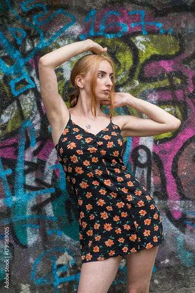 Obraz Blonde sensual woman with long legs posing confidently in front of a colorful graffiti wall, wearing a short floral dress. Urban fashion meets vibrant street art in this expressive portrait.