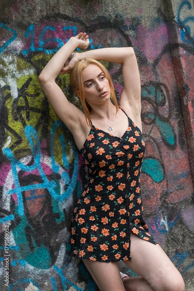 Obraz Blonde sensual woman with long legs posing confidently in front of a colorful graffiti wall, wearing a short floral dress. Urban fashion meets vibrant street art in this expressive portrait.