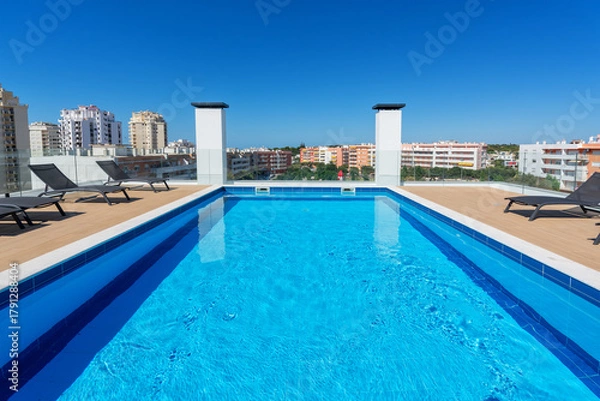 Fototapeta A vibrant rooftop pool sparkles under the sun, surrounded by lounge chairs, with a clear view of the cityscape and bright blue sky