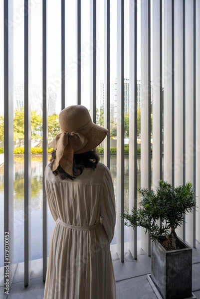 Fototapeta Woman in a Hat Behind a Modern Vertical Window with City View