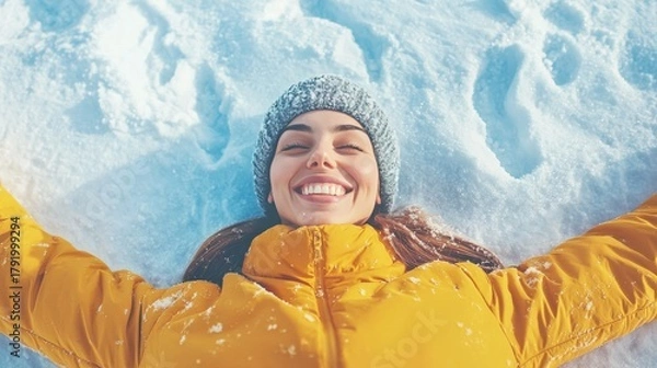 Fototapeta Joyful young woman in warm yellow jacket lying in fresh snow, smiling happily and enjoying a bright winter day outdoors