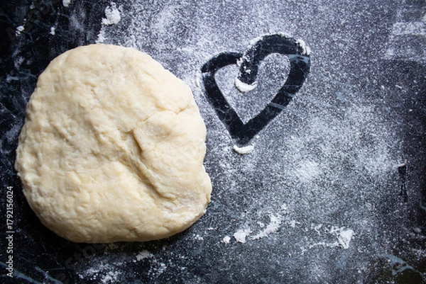 Fototapeta Raw dough on dark table with scattered flour and a heart drawn in it. Romantic rustic baking scene. Top view, homemade cooking concept, cozy kitchen atmosphere.