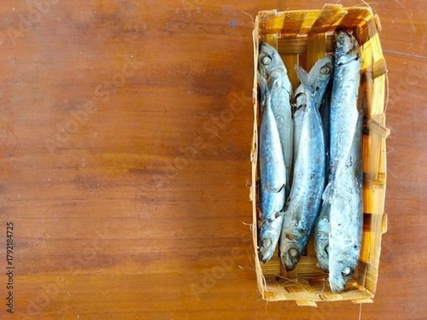 Fototapeta A woven basket holds several small, cooked or dried fish with silver and blue hues, resting on a warm wooden surface. Ikan pindang from a local market in Malang, Indonesia.