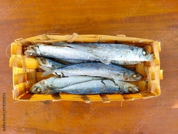 Fototapeta A woven basket holds several small, cooked or dried fish with silver and blue hues, resting on a warm wooden surface. Ikan pindang from a local market in Malang, Indonesia.