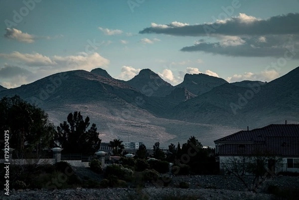 Fototapeta Scenic view of rugged mountain peaks under a moody sky with silhouetted houses and trees in the foreground at dusk