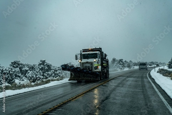 Fototapeta Snow plow truck clearing a wet road during a snowy winter day on a rural highway surrounded by snow-covered vegetation