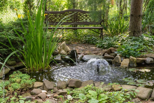 Fototapeta Serene garden pond with small waterfall, surrounded by rocks, lush greenery, and tall plants. Wooden bench is placed nearby, offering peaceful spot to relax amidst nature.