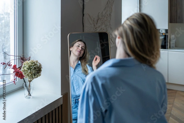 Fototapeta Confident contented middle aged woman enjoying looking at herself in mirror with self-acceptance, at home. Serene happy mature female embracing natural aging, feeling strong, beautiful. Beauty aging 