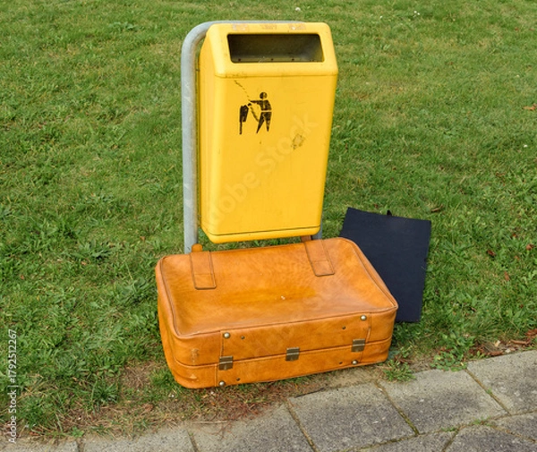 Obraz brown leather suitcase lies outside in front of a yellow trash bin on a lawn
