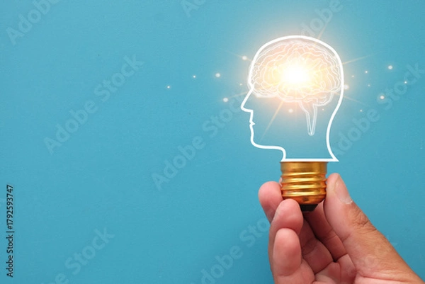 Fototapeta Creative idea and innovation concept. Hand holding lightbulb with glowing brain in head silhouette. Brainstorming, AI, knowledge, and new solution on blue background with copy space.