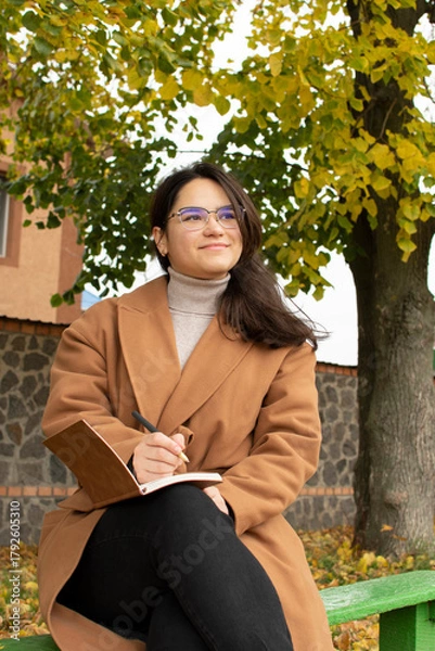 Fototapeta Young attractive woman in brown coat sitting on bench and writing in notebook	