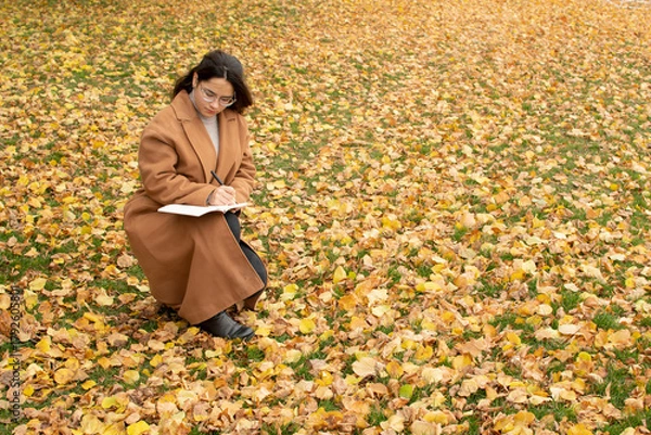 Fototapeta Young attractive woman in brown coat writing in notebook