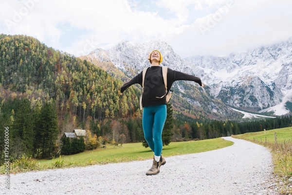 Fototapeta Young attractive woman hiking in nature. Beautiful happy girl stands on road looking at mountains view. Autumn or winter nature landscape. Photo of female having a walk with a backpack outdoors.