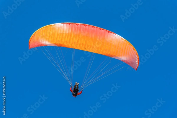 Fototapeta A single paraglider with a vibrant orange and red canopy is seen against a deep, clear blue sky during a sunny day flight.